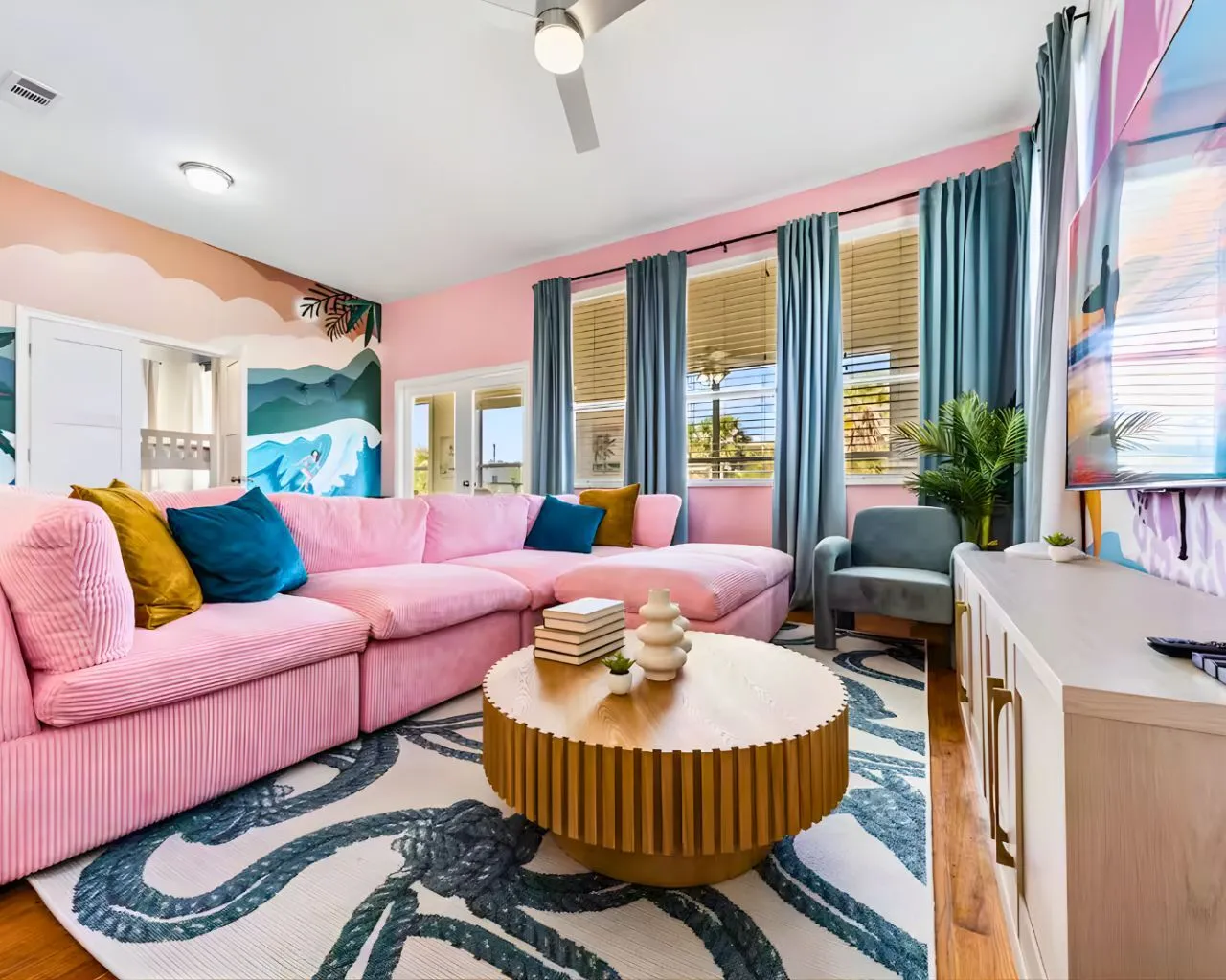 Bright living room with a pink sectional sofa, patterned rug, round wooden coffee table, blue curtains, and a mural wall with ocean waves.