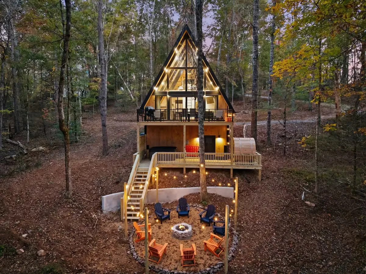 A-frame cabin in a wooded area at twilight with lit outdoor seating around a fire pit and balcony lights.
