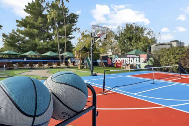Outdoor sports court with basketball hoop, two basketballs in foreground, and a blue and red court in the background.