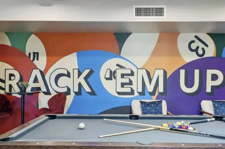 Indoor pool table with cues and balls set up, colorful wall mural behind reading 'RACK 'EM UP' with billiard ball designs.