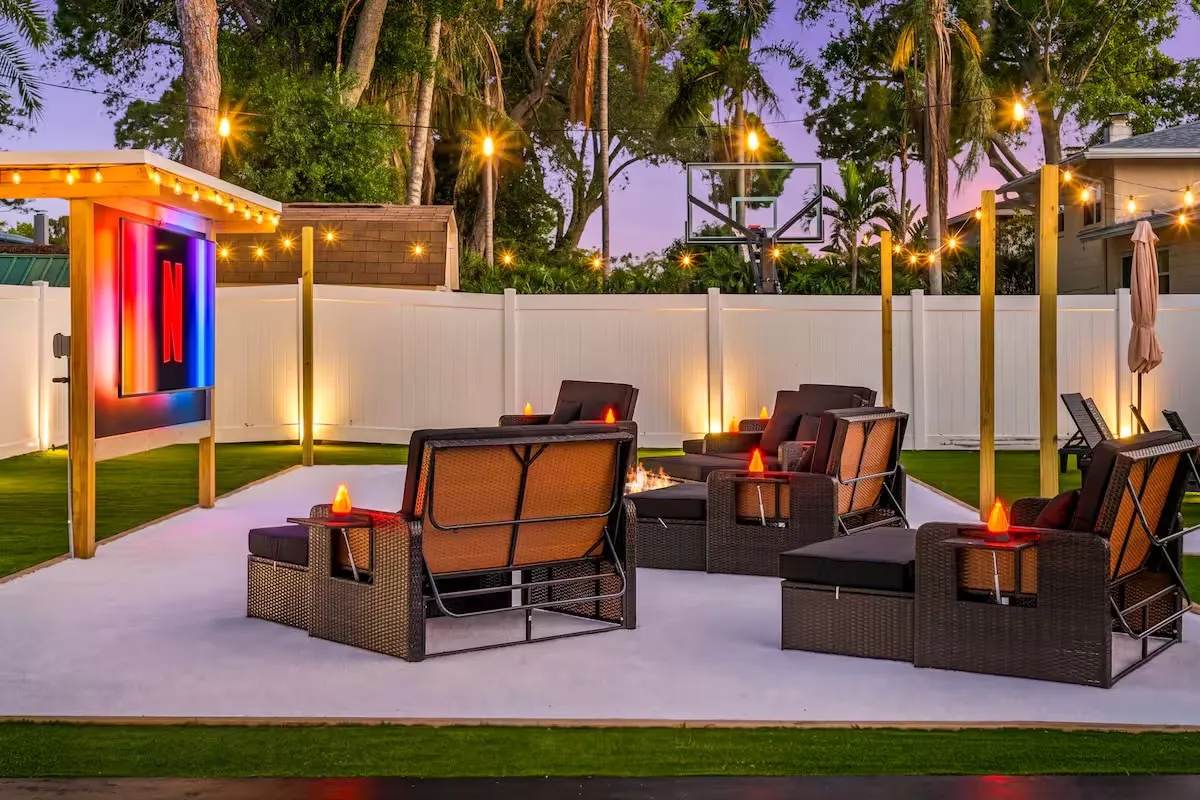 Outdoor patio with seating around a fire pit, string lights, a large TV screen.