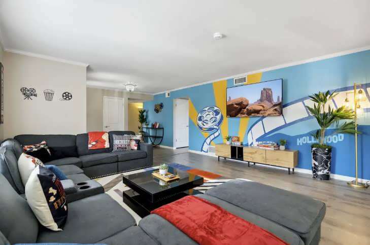 Modern living room with gray sectional sofa, colorful pillows, a black coffee table, and a blue Hollywood-themed mural with film reels on the wall behind a mounted flat-screen TV.